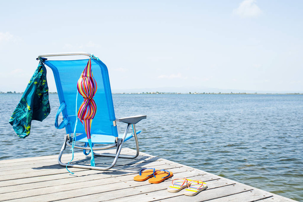 a colorful bikini and a pair of blue swimming trunks drying on a blue deck chair, and two pair of flip-flops on a wooden pier, next to the water - Fotografie, Obrázek