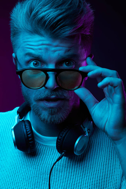 Neon light portrait of astonished man with headphones on neck, in sunglasses and white t-shirt.  - Фото, изображение