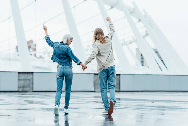 rear view of young couple holding hands and raising fists on street on cloudy day - Фото, изображение