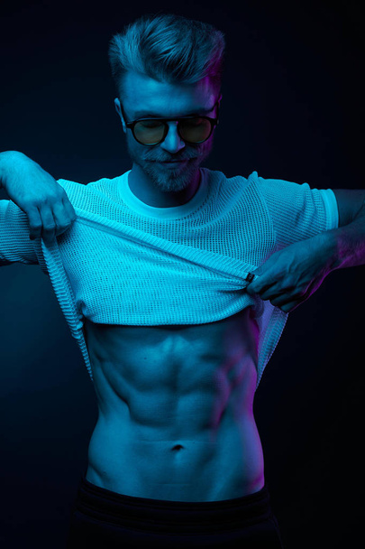Neon light portrait of sexy man in yellow glasses, pulling up t-shirt, showing abs - Photo, Image
