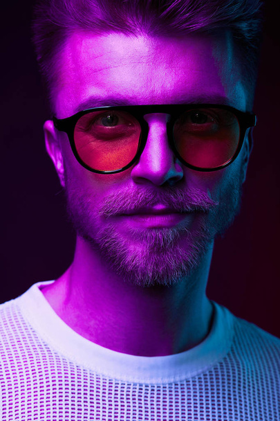 Neon light close-up portrait of serious man with mustaches and beard in orange sunglasses and white t-shirt  - Фото, изображение