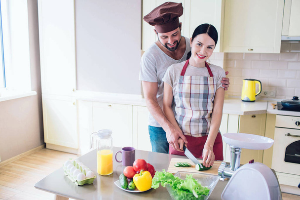 Beautiful couple stands at kitchen and wokr together. Girl looks at camera and smiles. She holds knife in one hand and cucumber in the other one. Guy helps her to cut vegetable. - 写真・画像