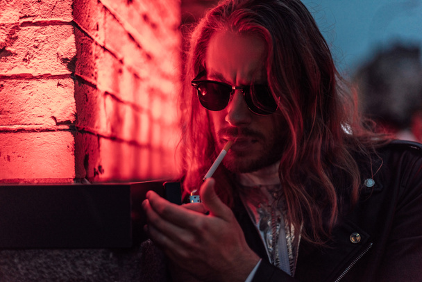 handsome young man in sunglasses and leather jacket smoking cigarette under red light on street - 写真・画像