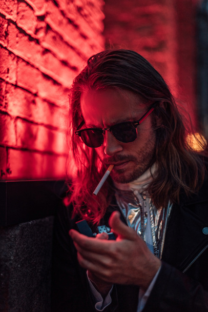 attractive young man in sunglasses and leather jacket smoking cigarette under red light on street - Φωτογραφία, εικόνα