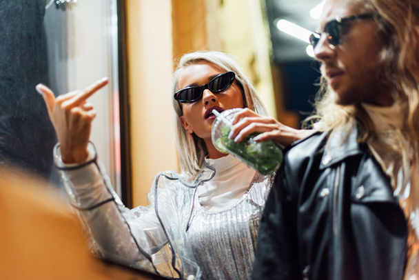 young stylish woman in sunglasses drinking mojito from plastic cup and showing middle finger at camera while spending time with boyfriend on street at night - Photo, Image
