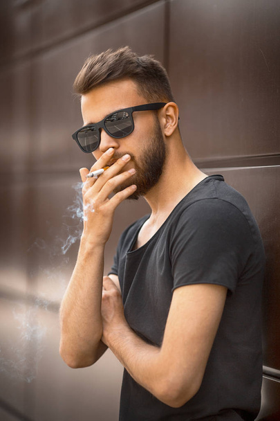 A young handsome white bearded man in sunglasses and black t-shirt smokes a cigarette in the street in the spring. Close up. - Фото, зображення