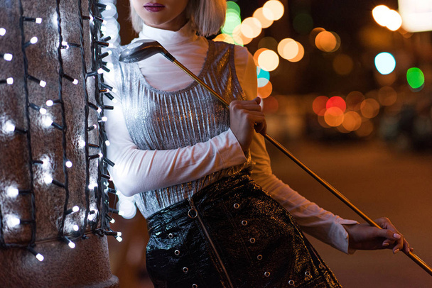 tiro recortado de la mujer joven con estilo club de golf apoyado en la pared con guirnalda blanca en la calle de la ciudad por la noche
 - Foto, imagen