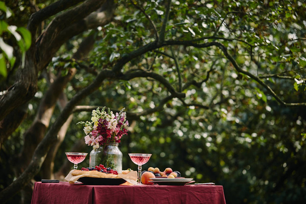 μπουκέτο με λουλούδια σε γυάλινο βάζο και wineglasses στο τραπέζι μέσα σε καταπράσινο κήπο - Φωτογραφία, εικόνα