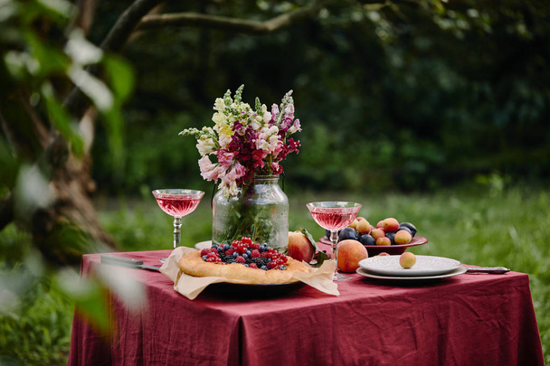 bouquet of flowers in glass jar, fruits and wineglasses on table in garden - Photo, Image