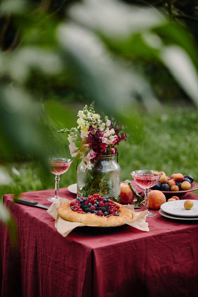 bouquet of flowers in glass jar, berries pie and wineglasses on table in garden - Zdjęcie, obraz
