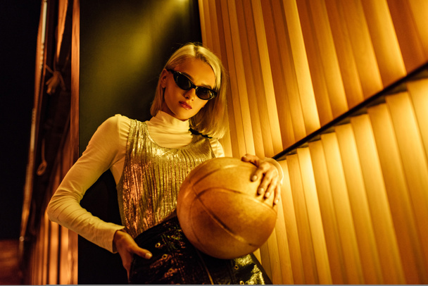 нижний вид молодой женщины в солнечных очках с золотистым глянцевым баскетбольным мячом на улице ночью под желтым светом
 - Фото, изображение