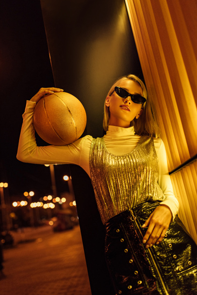 Onderaanzicht van aantrekkelijke vrouw in zonnebril met gouden glanzend basketbal bal op straat 's nachts onder geel licht - Foto, afbeelding