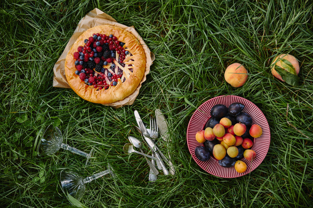 elevated view of pie with berries, plate with fruits and utensils on green grass - Valokuva, kuva