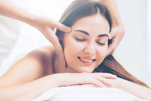 Zrelaksowana kobieta leżąca na łóżku spa na masaż twarzy i głowy zabieg spa przez masażystę w luksusowym kurorcie spa. Koncepcja odnowy biologicznej, stresu i odmłodzenia. - Zdjęcie, obraz