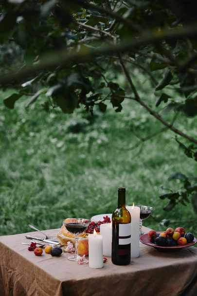 candles, wine bottle and fruits on table in garden for dinner - Foto, Imagen