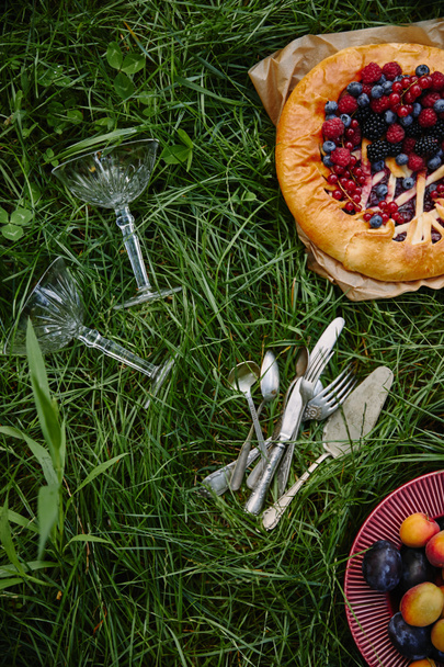 top view of pie with berries, plate with fruits and utensils on green grass - Photo, Image