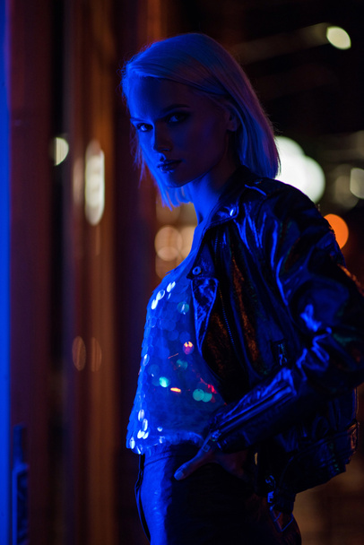 attractive young woman in glossy tank top and leather jacket on street at night under blue light - Foto, Imagen