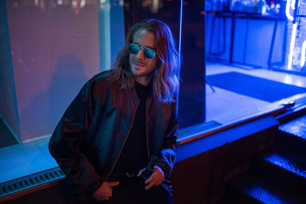 attractive young man in leather jacket and sunglasses with bottle of beer on street at night under blue light - Φωτογραφία, εικόνα