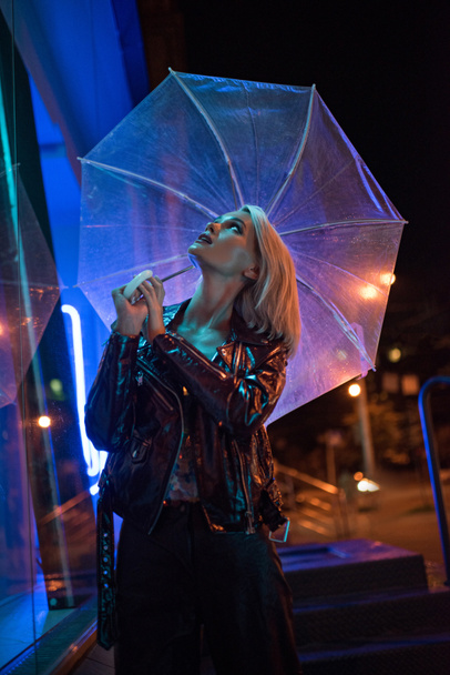 beautiful young woman with umbrella looking up on street at night under blue light - Zdjęcie, obraz