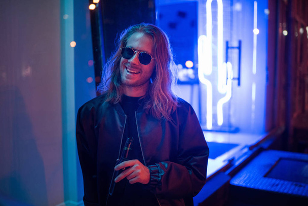 heureux jeune homme en veste en cuir et lunettes de soleil avec bouteille de bière dans la rue la nuit sous la lumière bleue
 - Photo, image