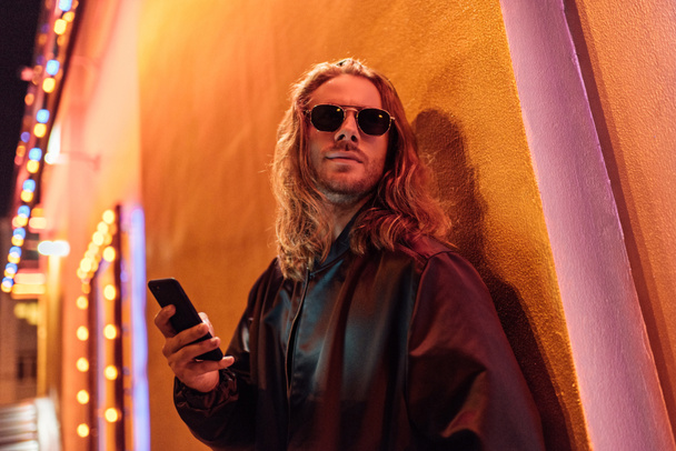 vista inferior do jovem atraente em jaqueta de couro e óculos de sol usando smartphone e olhando para a rua à noite sob luz amarela
 - Foto, Imagem
