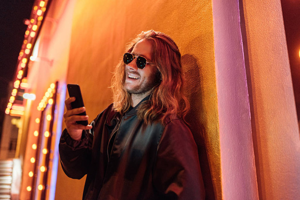 laughing young man in leather jacket and sunglasses using smartphone on street at night under yellow light - Photo, Image