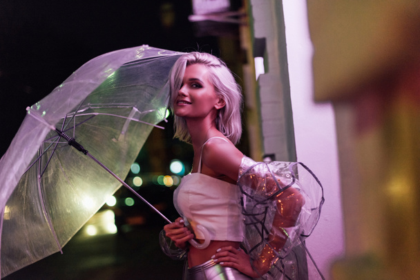 smiling young woman in transparent raincoat with umbrella on street at night under pink light - Photo, Image