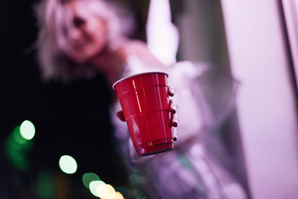 close-up shot of young woman holding red plastic cup on street at night under pink light - Photo, Image