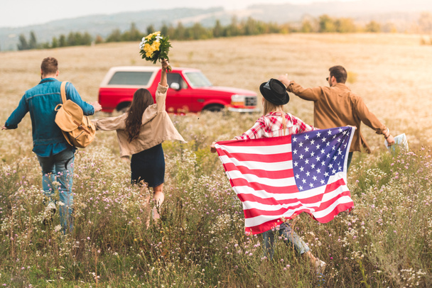visão traseira do grupo de jovens viajantes americanos com bandeira andando pelo campo de flores durante a viagem de carro
 - Foto, Imagem