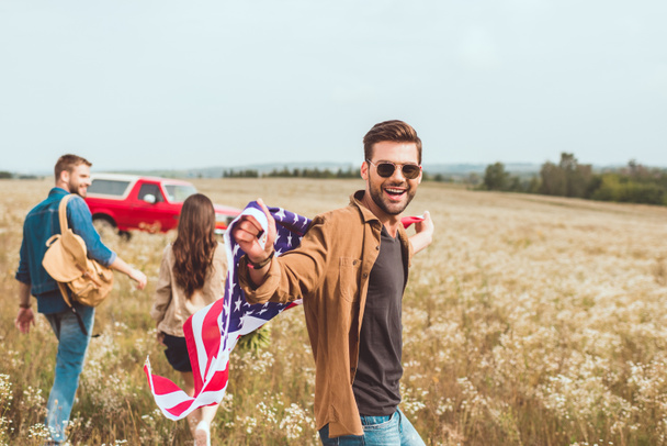 handsome young man with united states flag in flower field with friend during car trip - Photo, Image