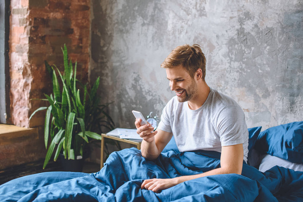 sourire jeune homme vérifier smartphone dans le lit à la maison
  - Photo, image