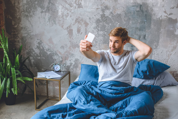 handsome young man taking selfie on smartphone in bed at home - Fotó, kép