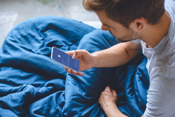 selective focus of young man in bed using smartphone with facebook on screen  - Fotografie, Obrázek