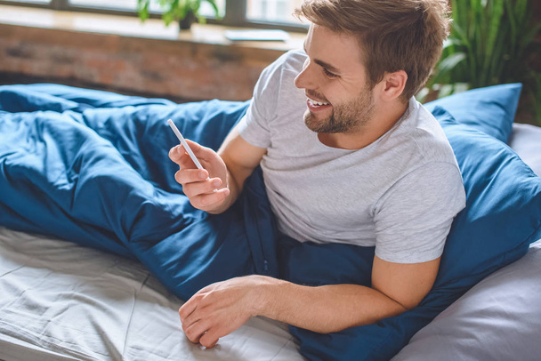 χαμογελαστός νεαρός άνδρας τον έλεγχο smartphone στο κρεβάτι στο σπίτι - Φωτογραφία, εικόνα