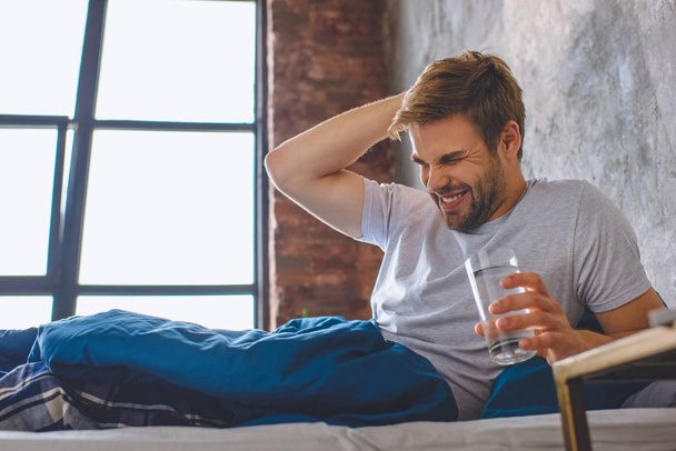 young man with headache holding glass of water in bed at home - Photo, image