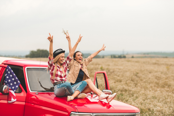 schöne junge Freundinnen sitzen mit erhobenen Händen auf der Motorhaube und zeigen Friedenszeichen - Foto, Bild