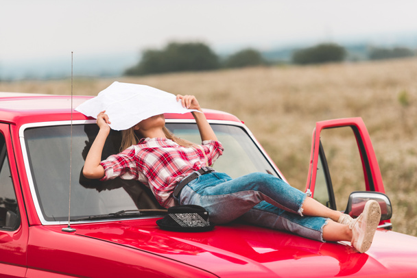 young woman lying on hood of vintage red truck in field and covering face with map in field - Photo, Image