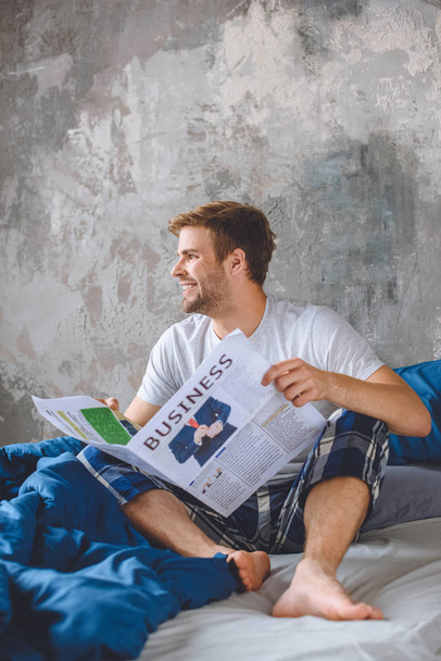 smiling young man reading business newspaper in bed during morning time at home - 写真・画像
