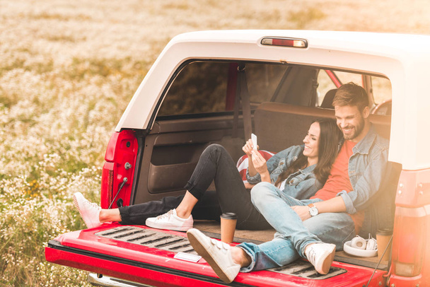 beautiful young couple using smartphone while relaxing in car trunk - Foto, imagen