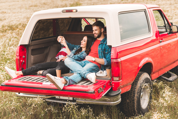 gyönyörű fiatal pár véve selfie smartphone, míg pihen a autó törzs - Fotó, kép