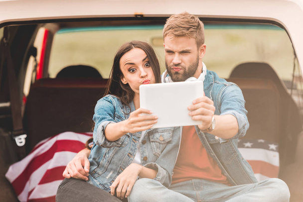 beautiful young couple making selfie with tablet while sitting in car trunk during trip - Foto, immagini