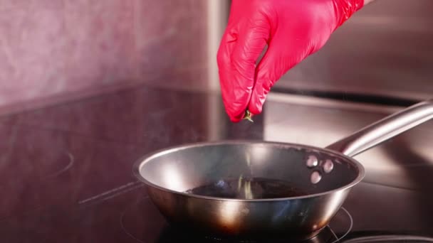 Chef-koks hand giet rozemarijn op een hete koekenpan op commerciële keuken. Olie in de pan is sissende en spuiten, close-up in slow motion. - Video