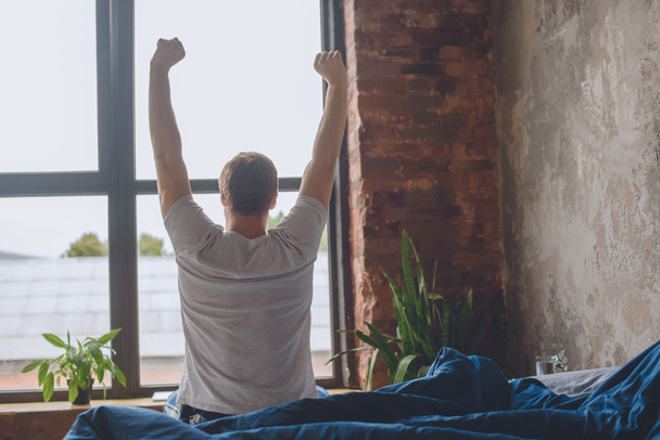 вид сзади на молодого человека, сидящего на кровати с поднятыми руками в утреннее время дома
  - Фото, изображение