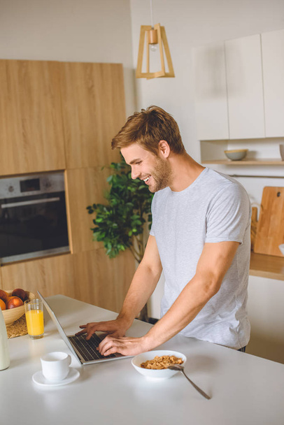 smiling young man using laptop at kitchen table with breakfast  - Foto, Imagem