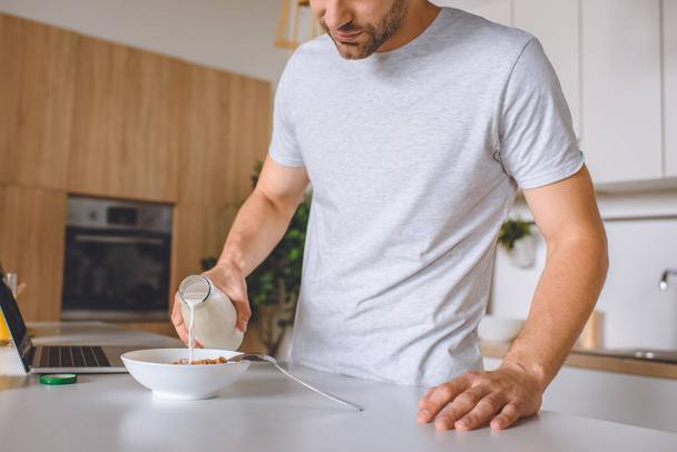 cropped image of man pouring milk into plate with flakes at kitchen table with laptop  - Foto, immagini