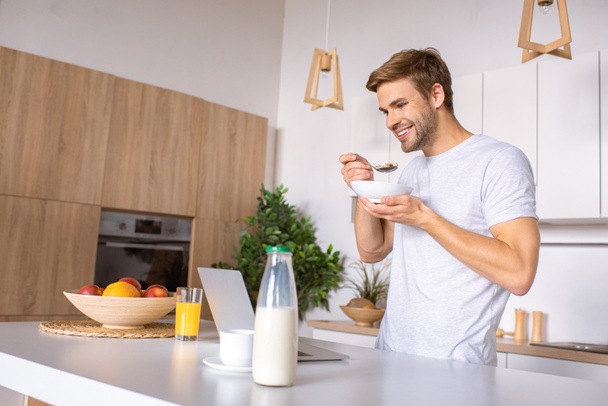 smiling man eating flakes with milk on breakfast and looking at laptop screen at kitchen  - Foto, imagen