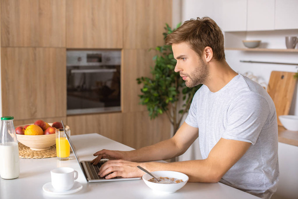 joven enfocado utilizando el ordenador portátil en la mesa de la cocina con desayuno
  - Foto, Imagen
