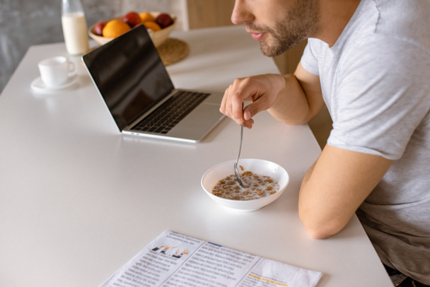 περικομμένη εικόνα άνθρωπος τρώει νιφάδες με γάλα για πρωινό στο τραπέζι της κουζίνας με φορητό υπολογιστή  - Φωτογραφία, εικόνα