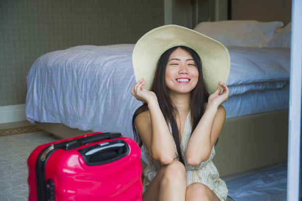 lifestyle natural portrait of young happy and beautiful Asian Korean tourist woman with travel suitcase arriving at hotel sitting on room floor excited and playful in Summer hat having holidays - Foto, Imagem