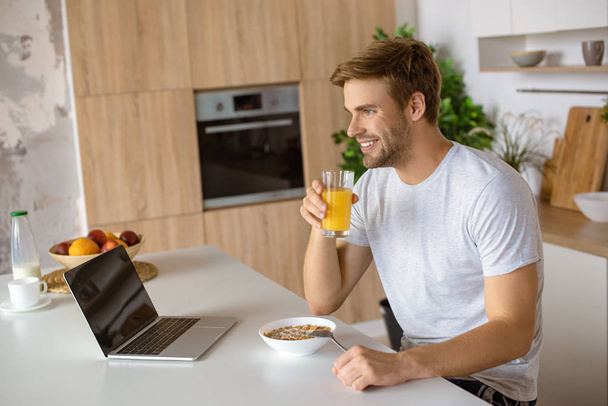 hymyilevä mies juo tuoretta mehua pöydässä levy hiutaleita ja kannettava tietokone keittiössä
  - Valokuva, kuva